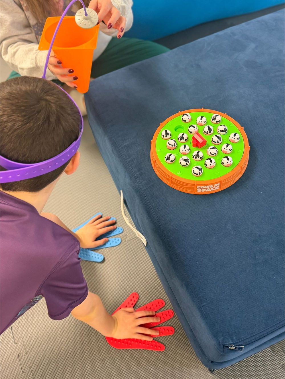 Child on hands and knees playing Cows in Space at NeuroNest Pediatric Therapy, with hand markers on the floor